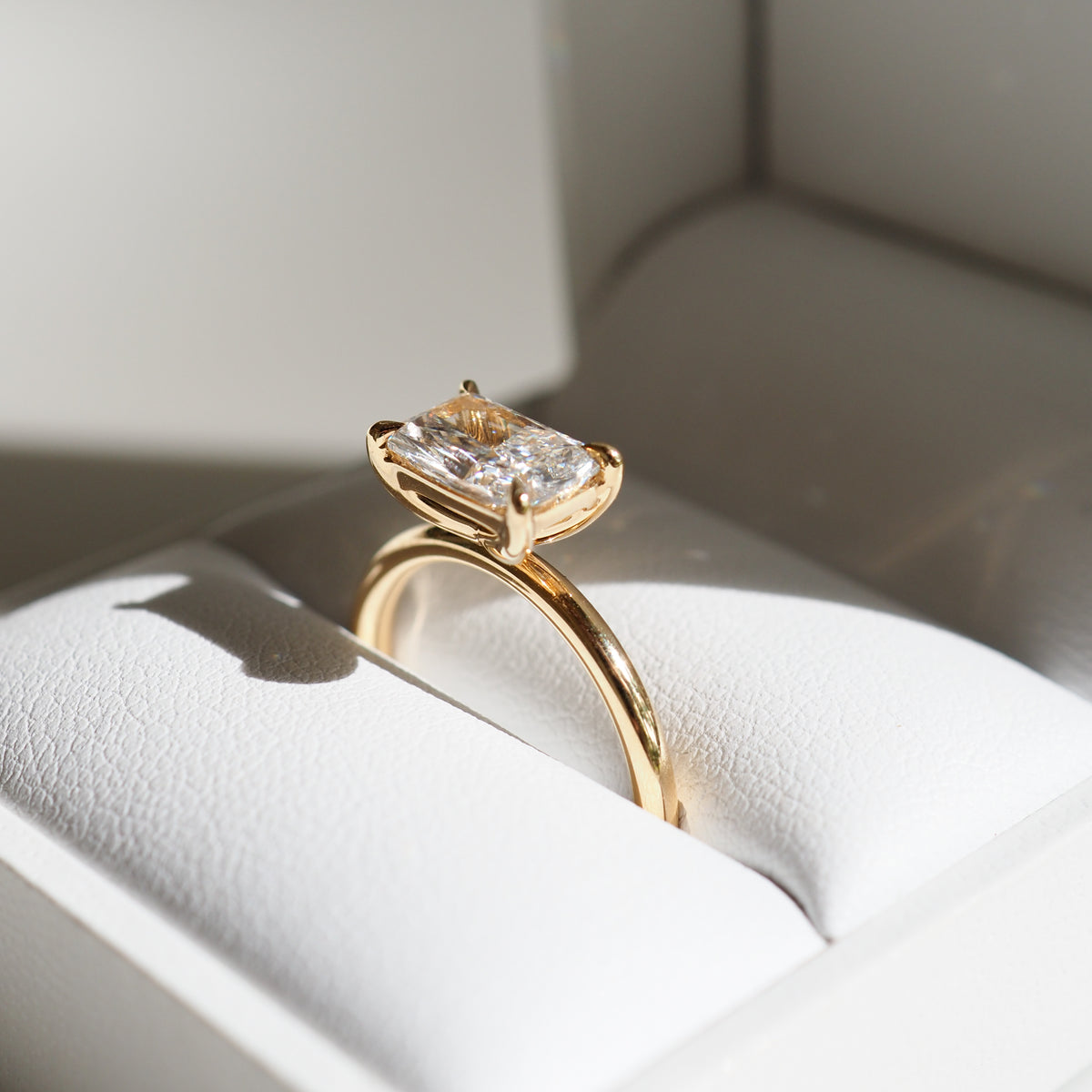Elegant gold ring featuring a radiant-cut diamond, displayed in a luxurious white jewellery box under soft lighting.