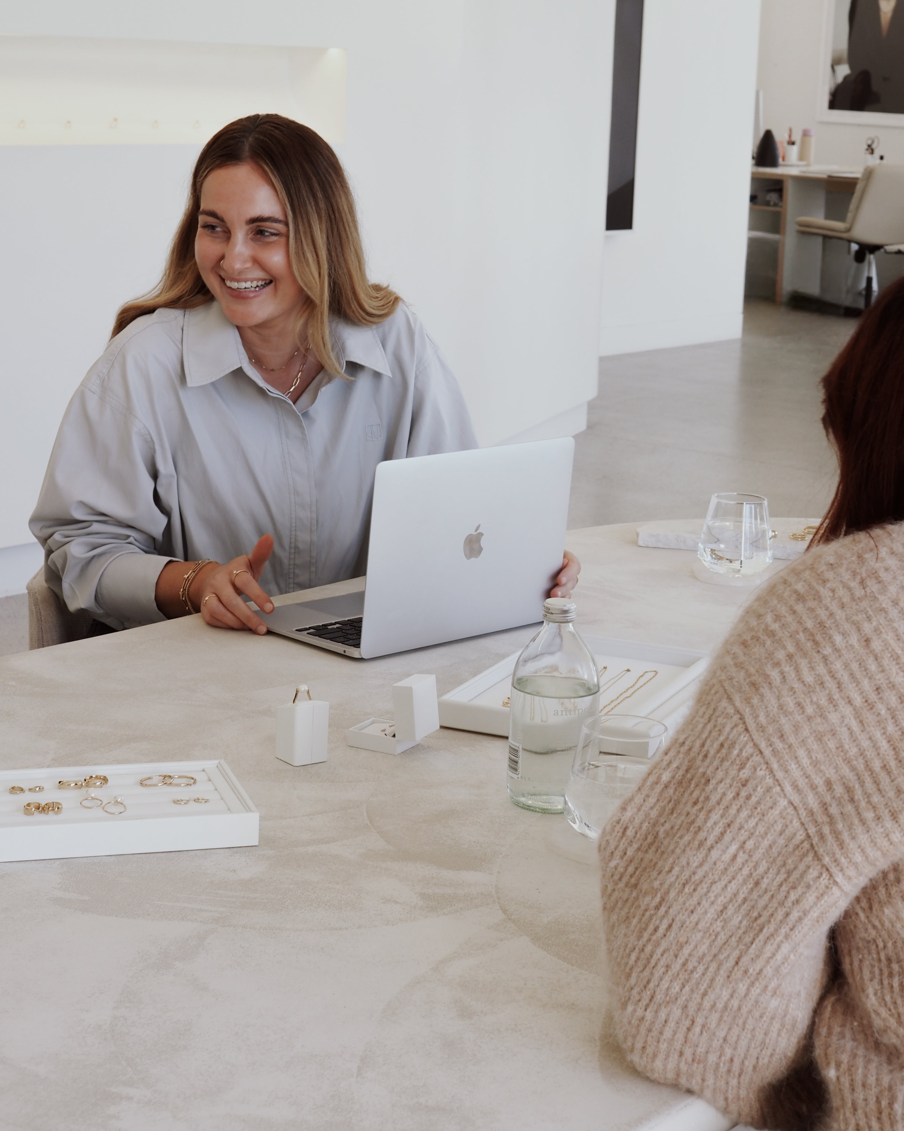 Two women at a luxury jewellery meeting. One uses a laptop, surrounded by a tray of gold jewellery, glassware, and decor on a sleek table.