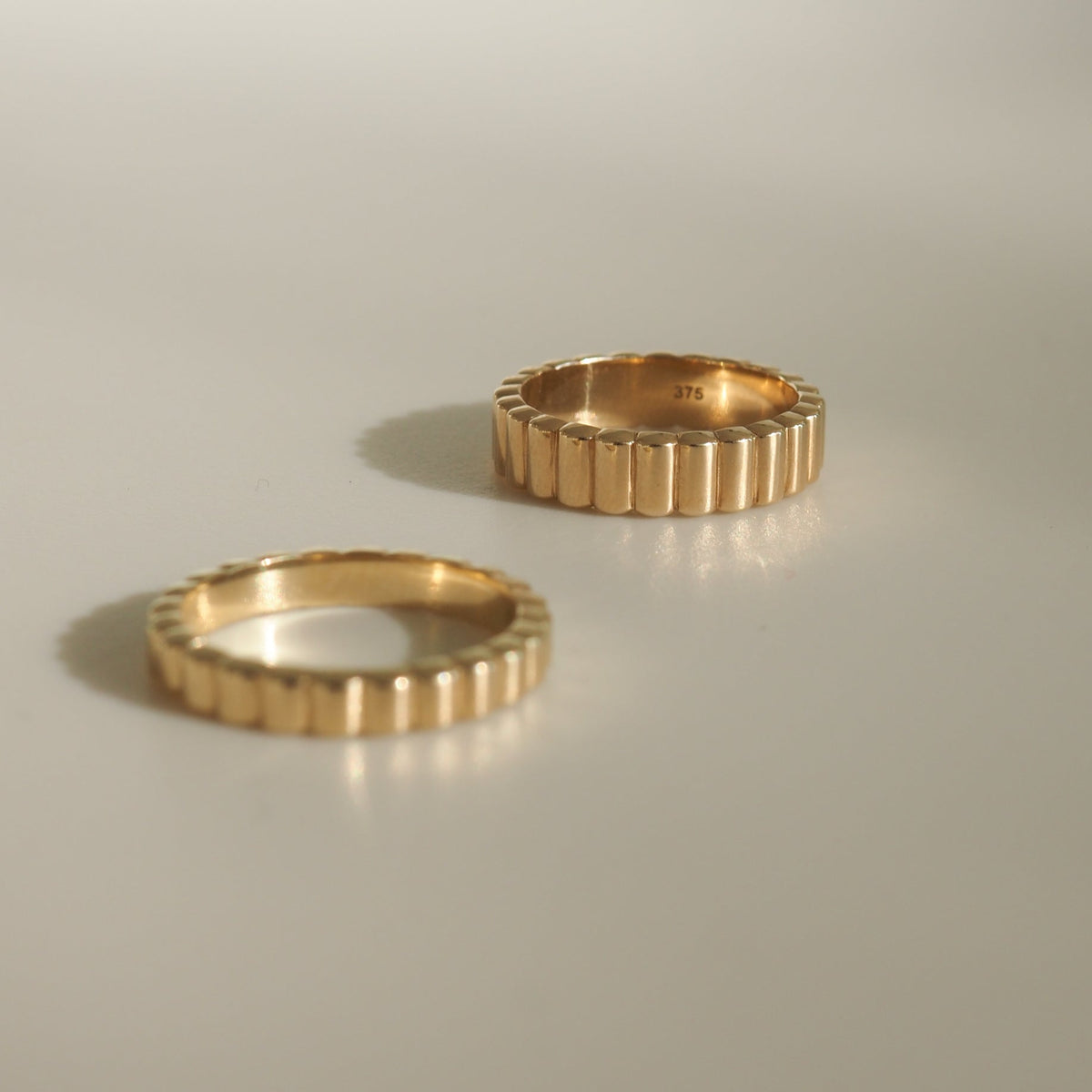 Two ribbed gold rings on a light surface, one standing upright showing "375" hallmark inside. Luxurious jewellery, minimalistic design.