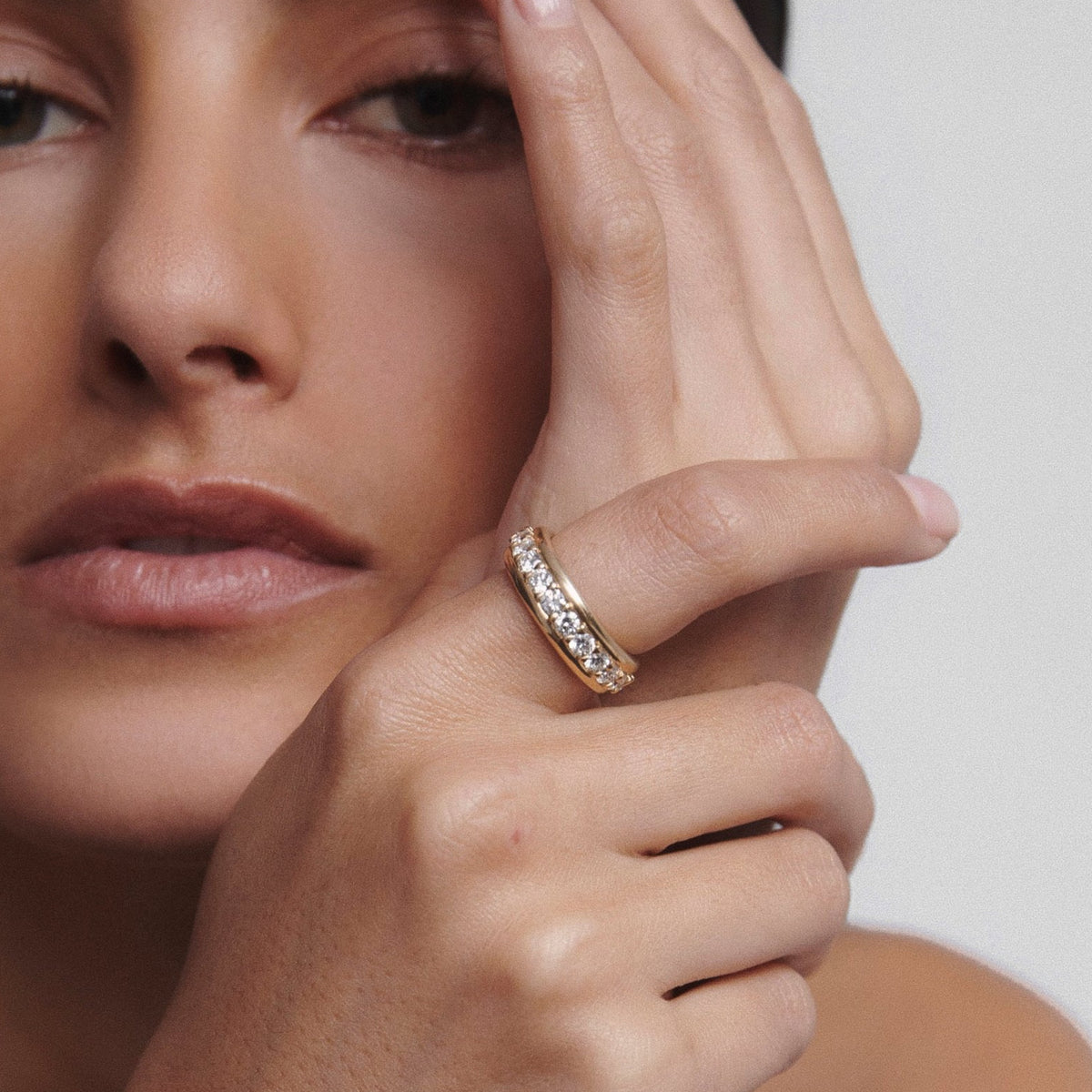 Woman with minimal makeup showcasing a gold diamond ring on her finger, hand resting on her face, set against a plain background.