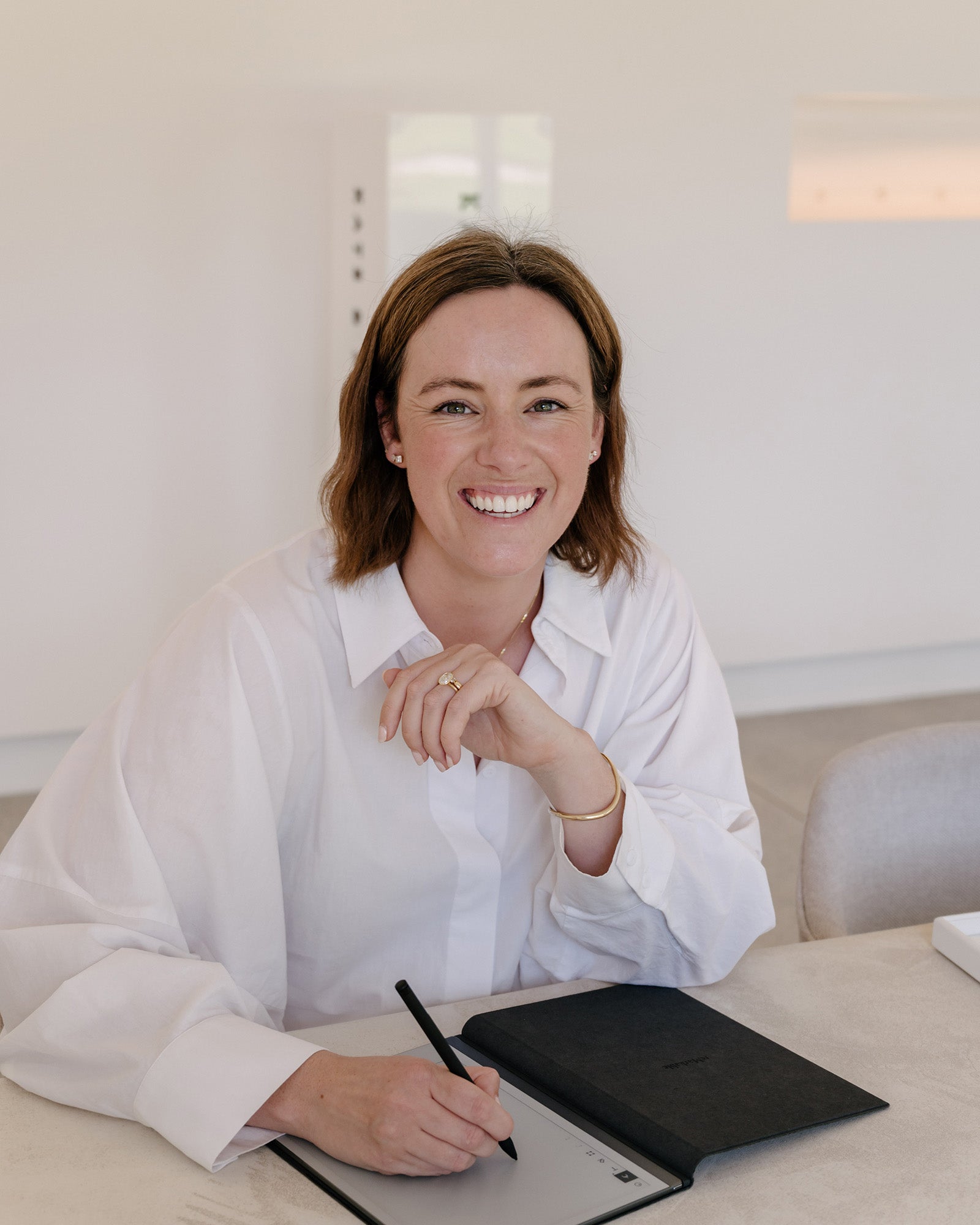 Smiling woman in white blouse writing in a notebook at a table, showcasing luxury jewellery in a minimalist setting.
