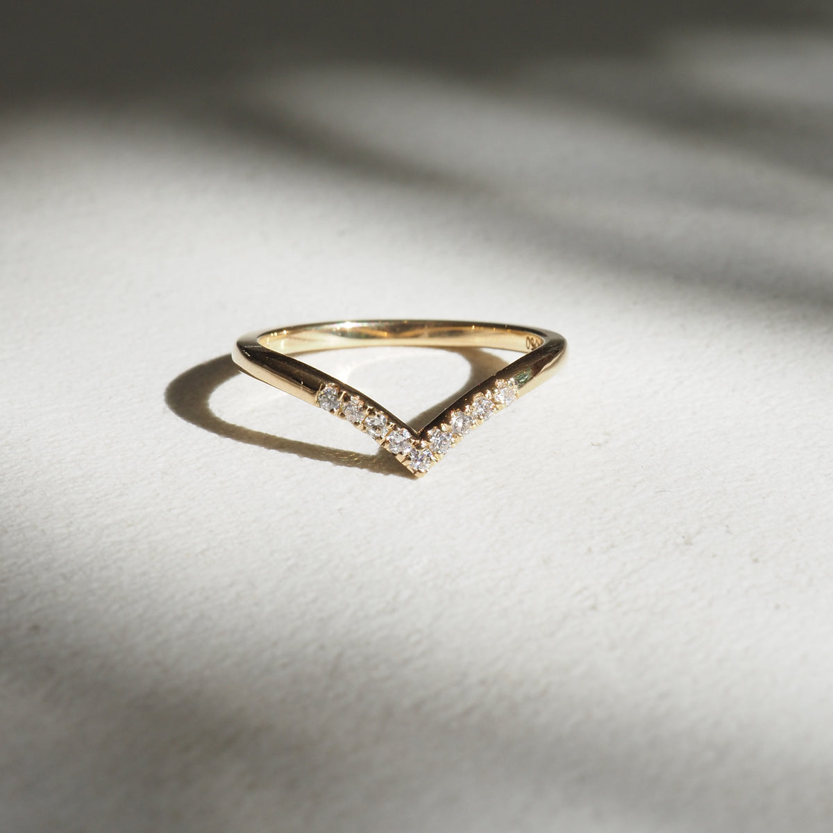 Gold V-shaped ring with small diamonds set along the top, casting a shadow on a light surface; elegant luxury jewellery piece.