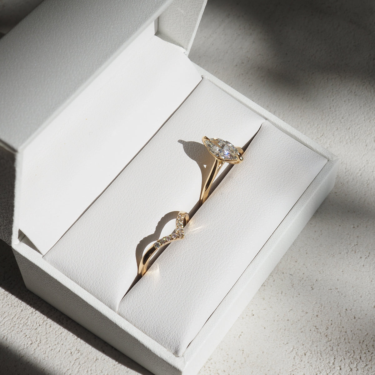 Gold engagement ring with marquise diamond in a white jewellery box. Sunlight casts soft shadows on elegant design.