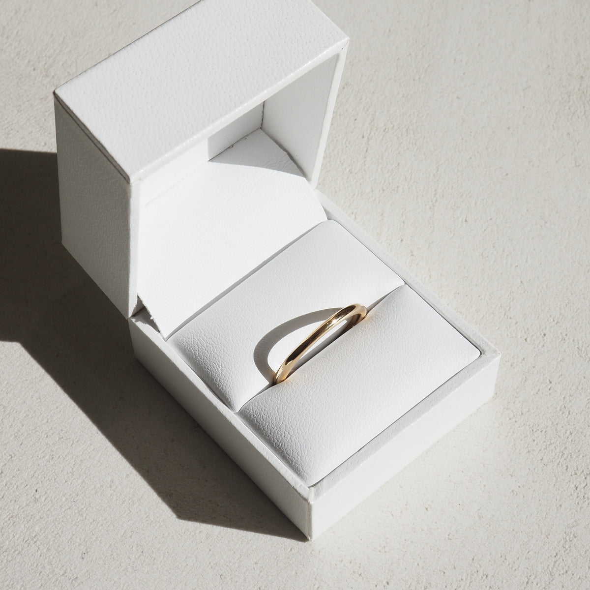 Elegant gold ring in a white leather jewellery box, sunlight casting a shadow, emphasising its luxury and minimalist design.