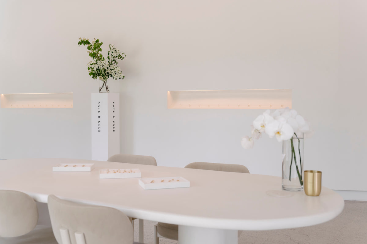 Minimalist luxury jewellery showroom with white table, beige chairs, floral centrepiece, and display boxes. Elegant, understated design.