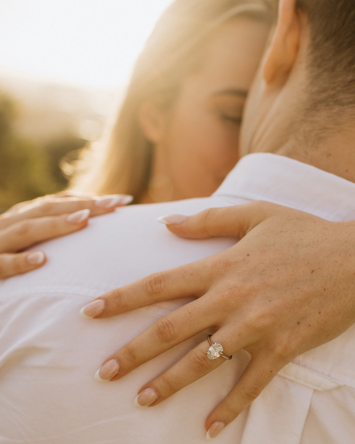 Couple embraces in sunlight, showcasing a luxurious diamond engagement ring against a white shirt. Romantic and elegant jewellery moment.