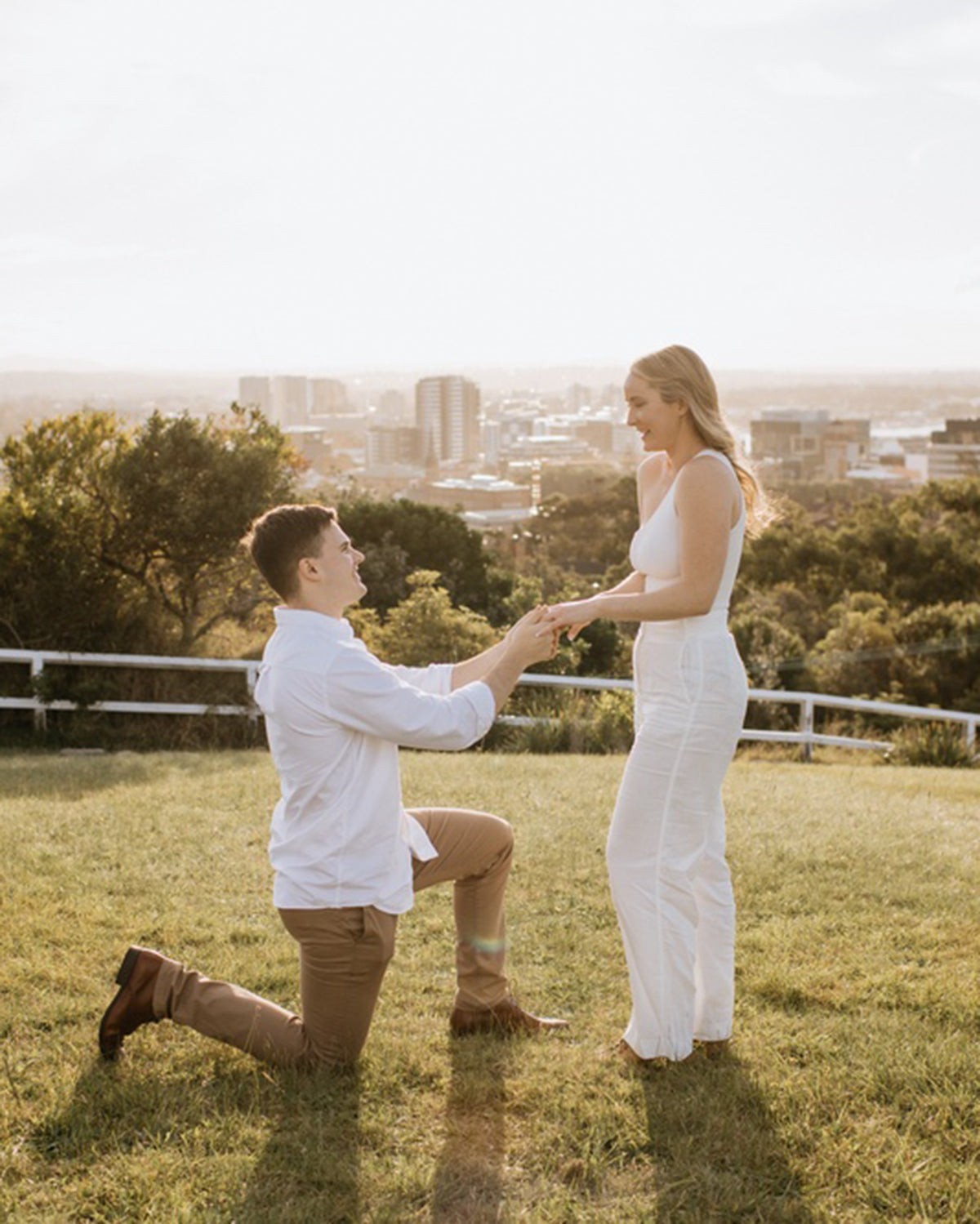 Man kneeling in proposal with engagement ring, woman standing, cityscape backdrop. Romantic, outdoor setting capturing sunset moment.