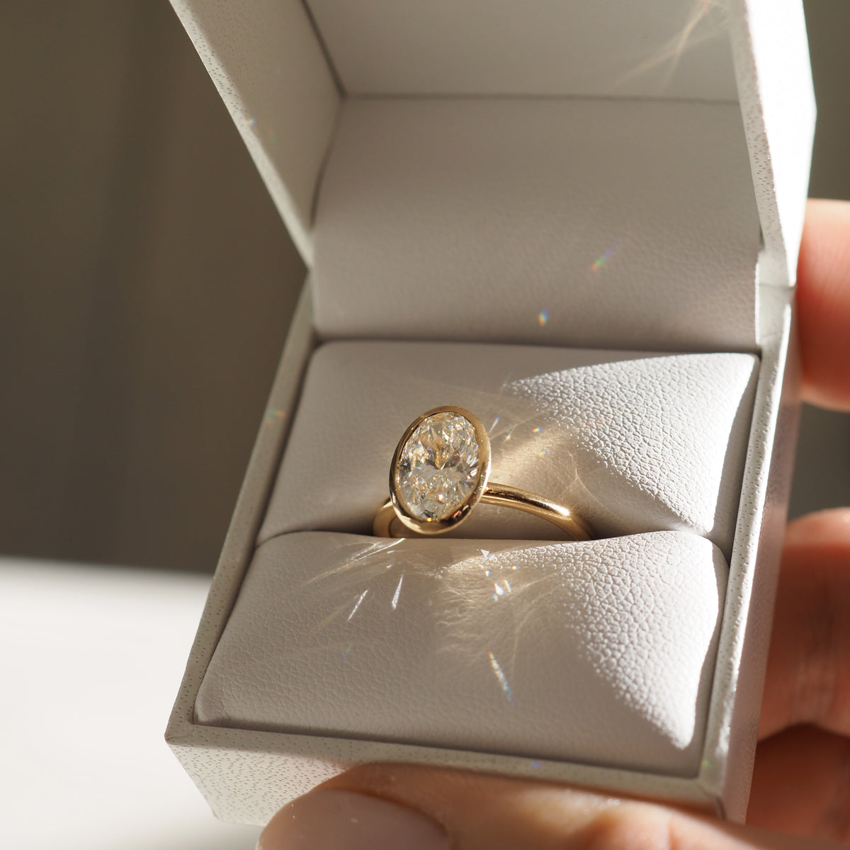 A gold band with an oval diamond ring displayed in an open white jewellery box, sparkling under soft light. Perfect for luxury engagements.