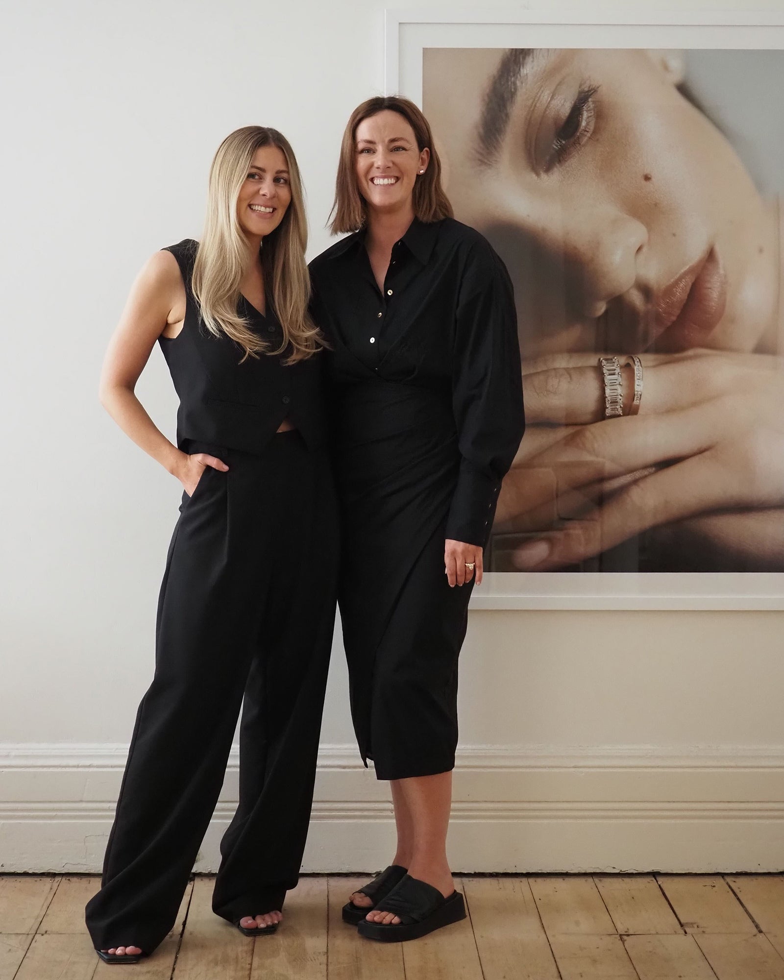 Two women in black outfits stand beside each other, smiling. Behind them is a large photograph of jewellery on a woman's hand.