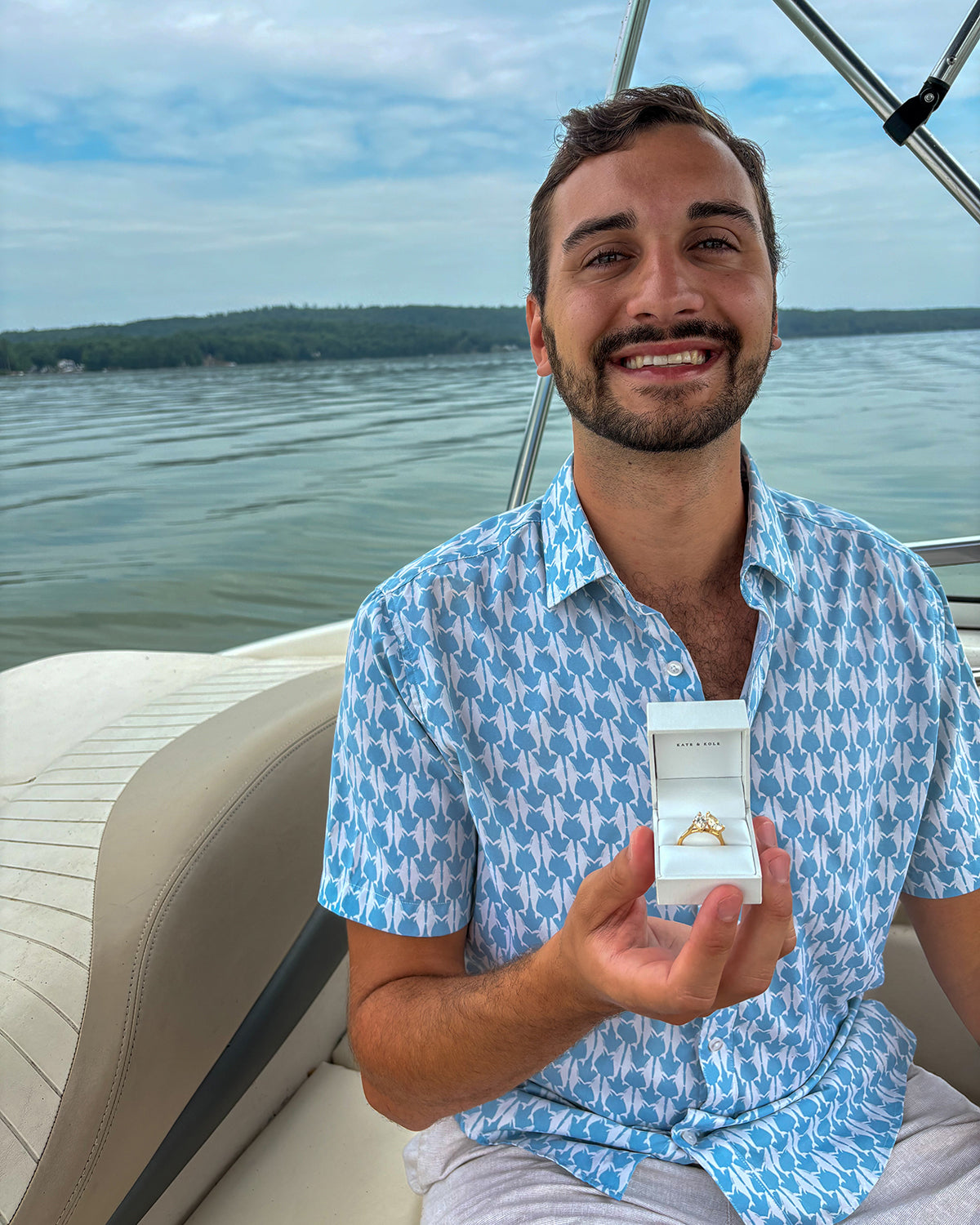 Man smiling on a boat, holding a white jewellery box with a gold ring. Background features calm water and distant trees.