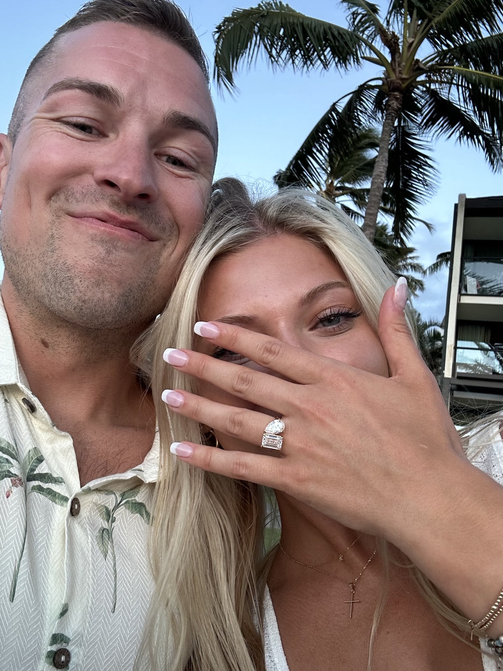Couple outdoors, woman showing off a luxury diamond engagement ring, palm trees in background.