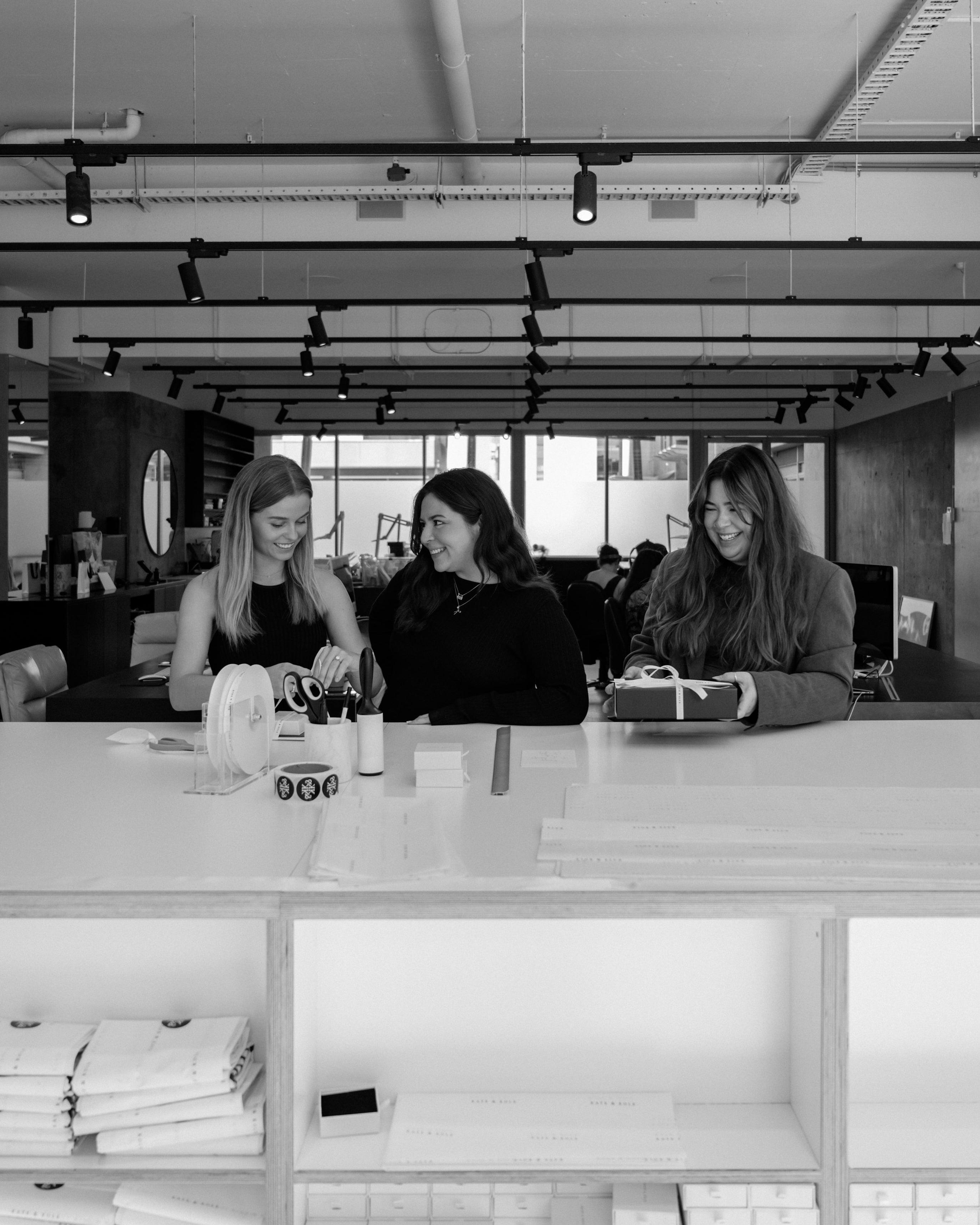 Three women are sitting at a modern jewellery showroom table, smiling and talking, with elegant decor and gift boxes around.