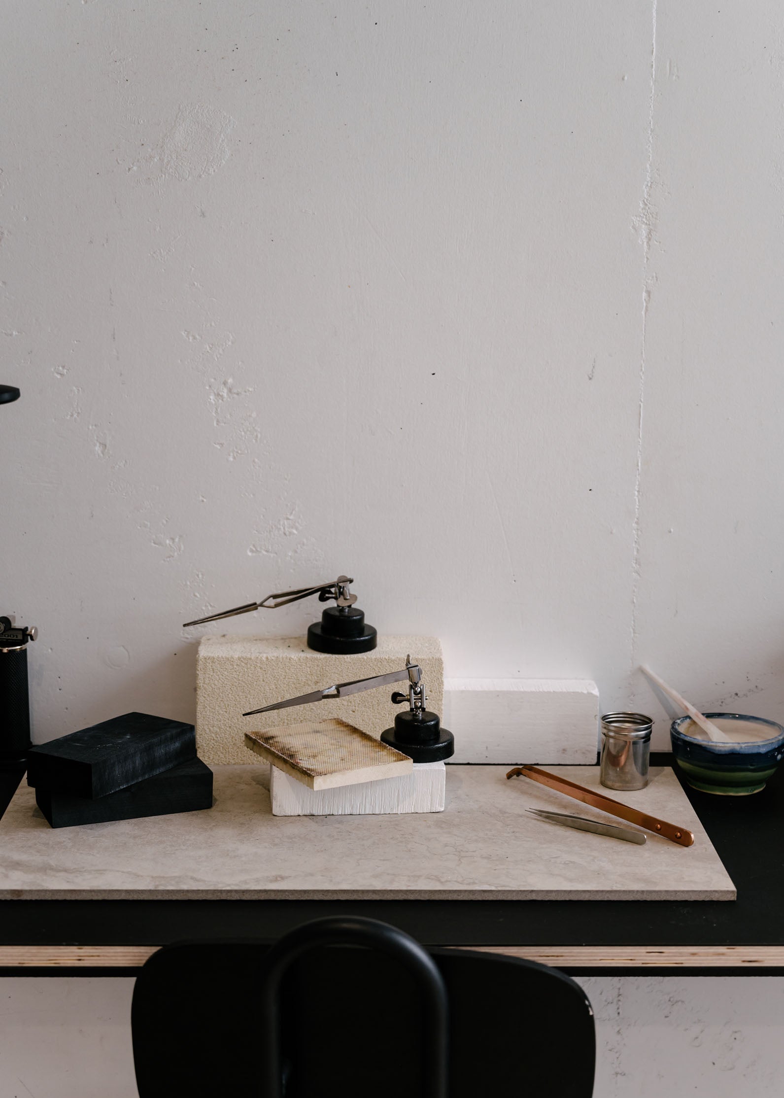 A minimalist jewellery workbench with white bricks, black tools, and small containers placed on a sleek black surface against a plain wall.