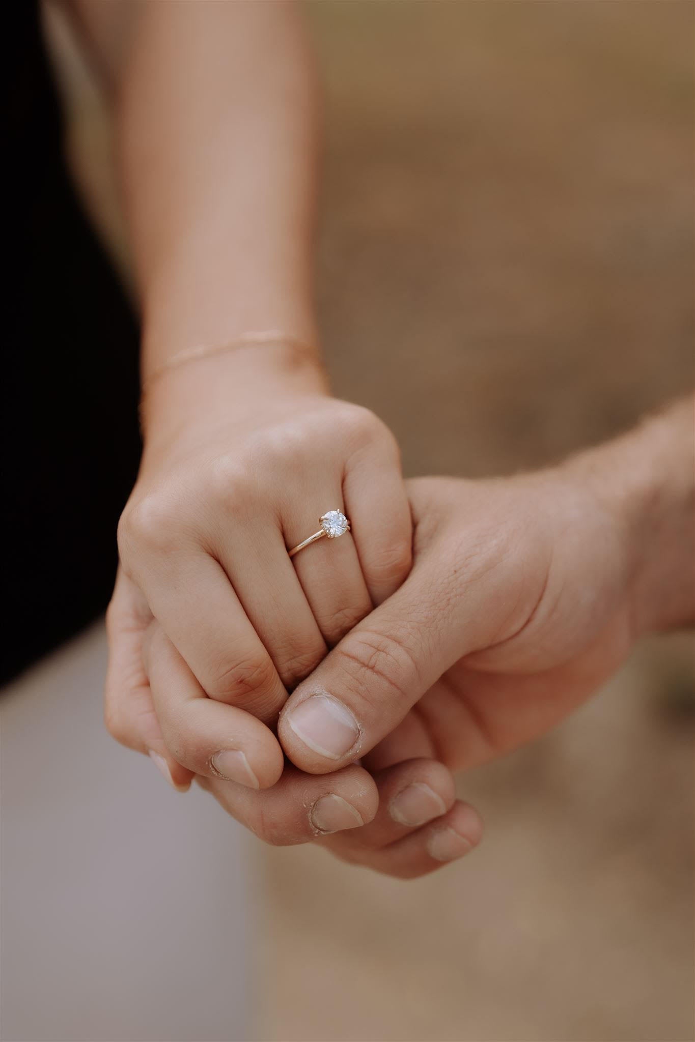 Hands holding, featuring a luxurious diamond engagement ring on a woman's finger. The background is softly blurred, enhancing focus.