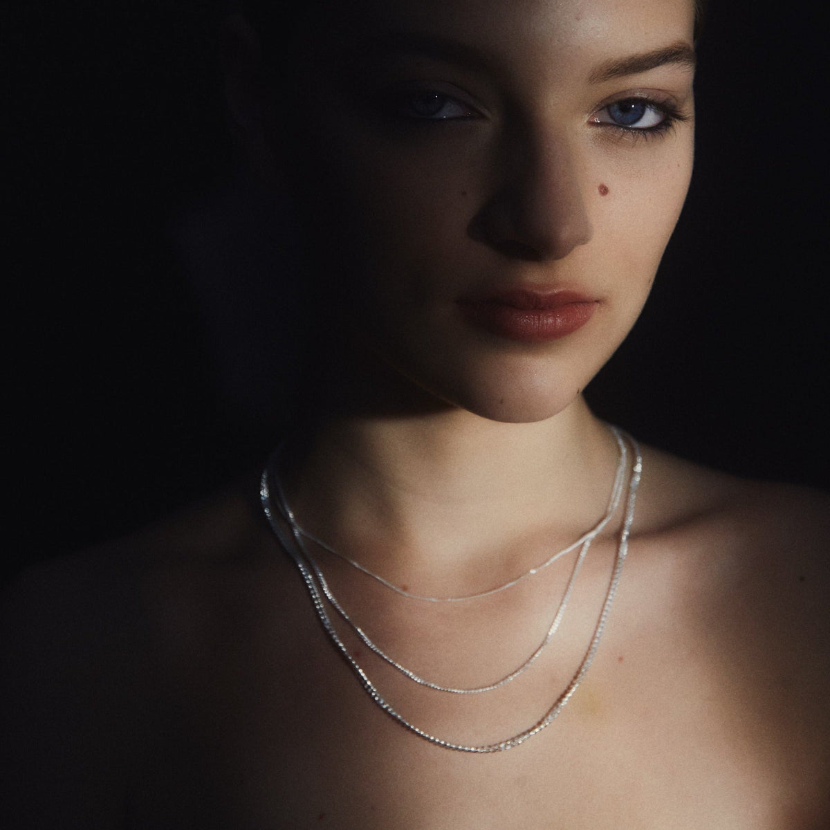 Woman wearing delicate layered silver necklaces in soft, moody lighting. Highlight on luxury jewellery with an artistic touch.